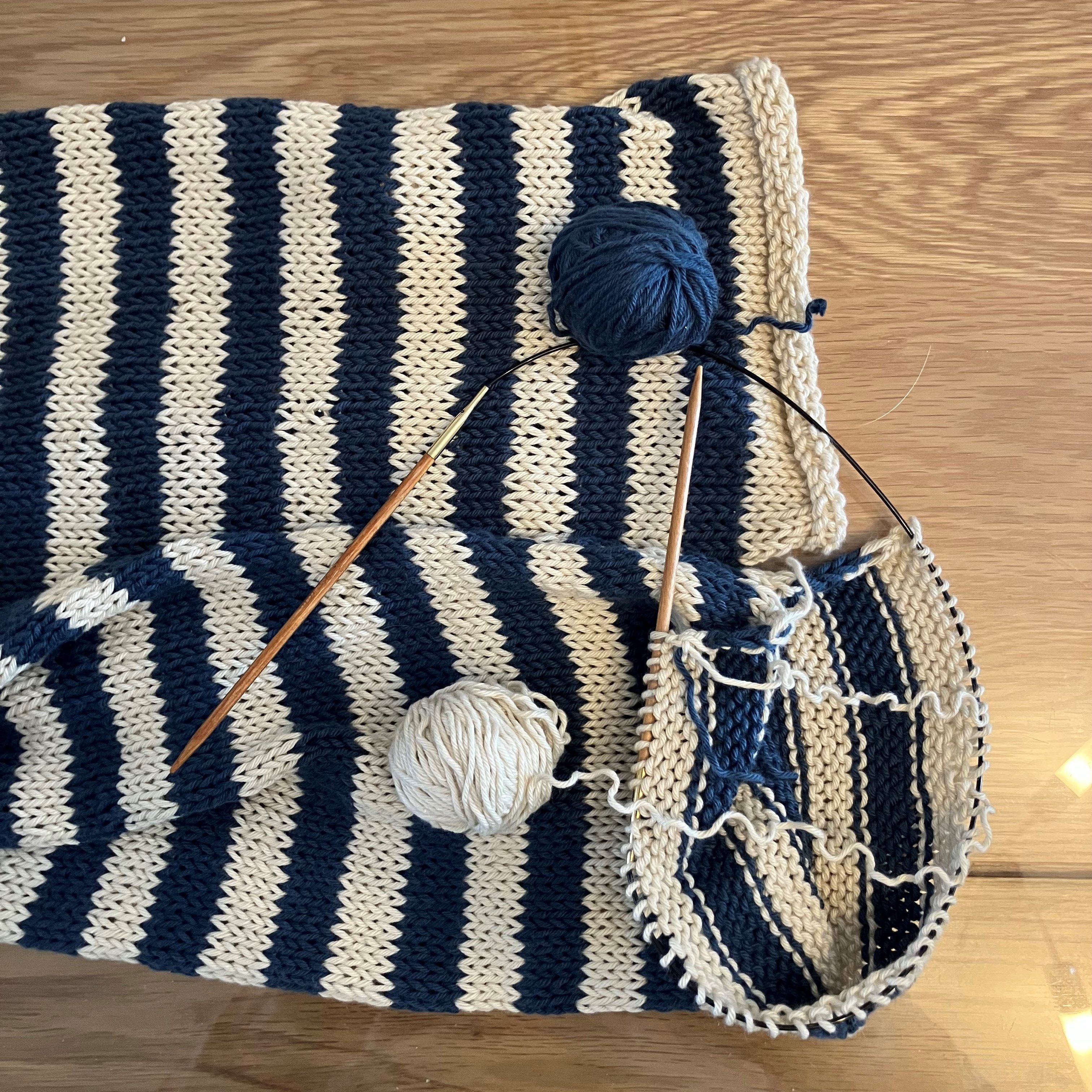 Close-up of a knitted project featuring a striped pattern in navy blue and cream, with knitting needles and two balls of yarn on a wooden surface.