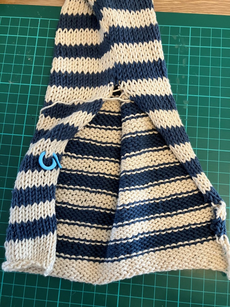 A close-up of a knitted fabric featuring alternating stripes of navy blue and cream, with a stitch marker placed in the fabric.