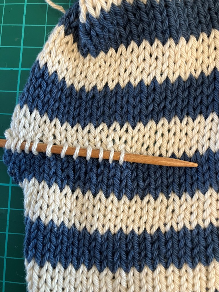 Close-up of a knitting project with navy and cream striped fabric, showing a wooden knitting needle inserted into the stitches.