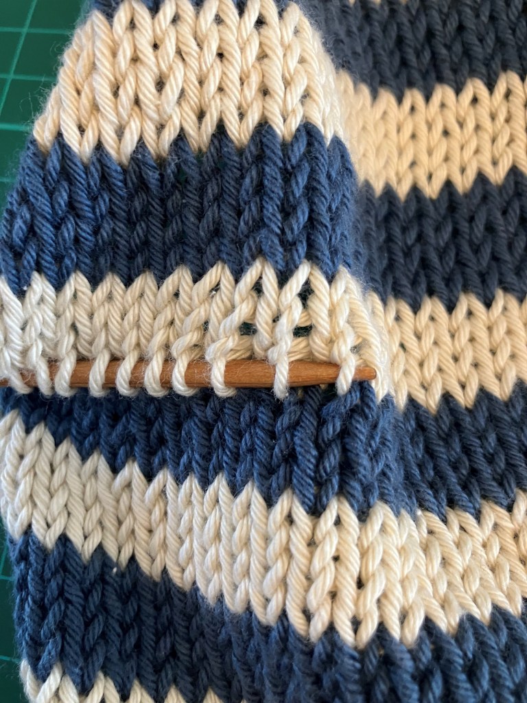 Close-up of a knitting project featuring blue and cream stripes, with a wooden knitting needle inserted into the fabric.