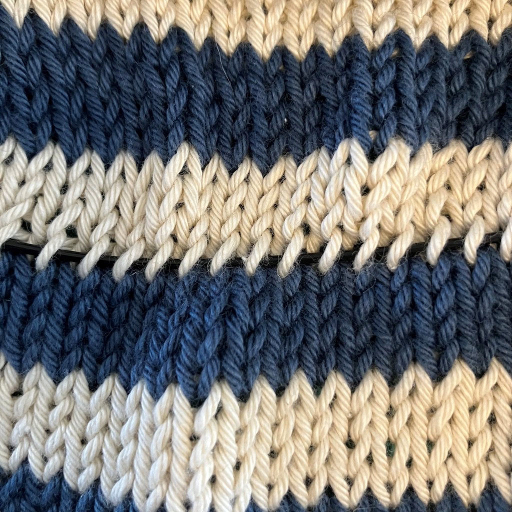 Close-up of knitted fabric in alternating stripes of blue and cream, showing a stitch marker threaded through the yarn.