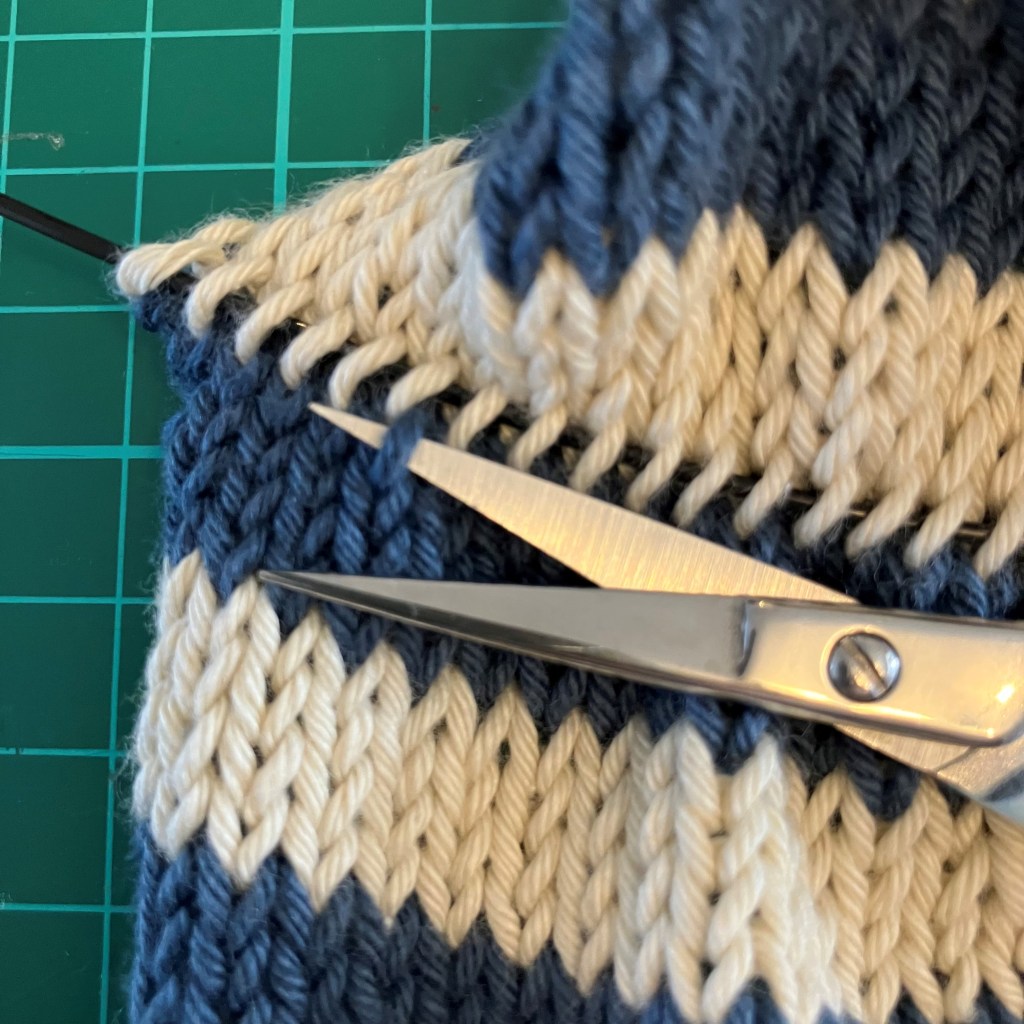 A close-up image showing scissors positioned near a knitted fabric with blue and cream stripes, indicating a knitting technique step.
