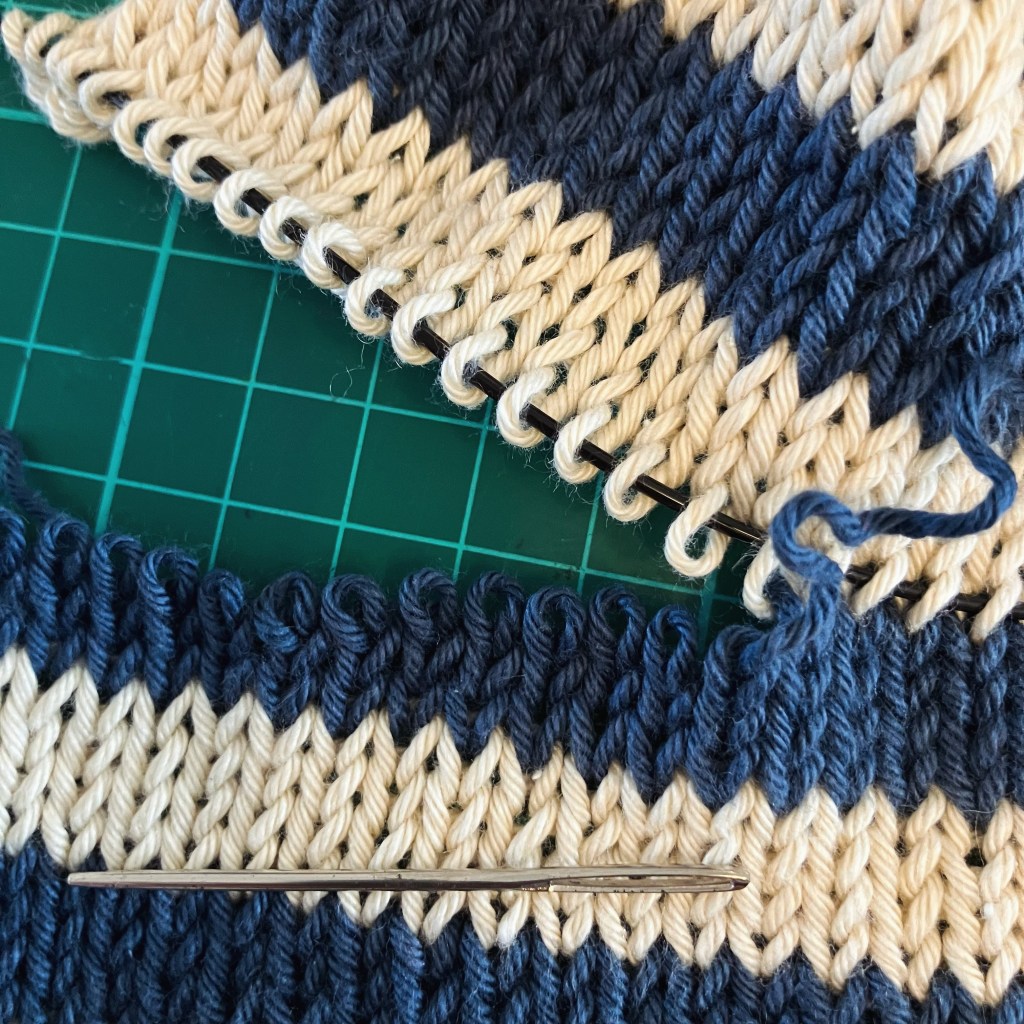 Close-up of knitted fabric showing blue and cream stripes, with a wool needle positioned on a green cutting mat.